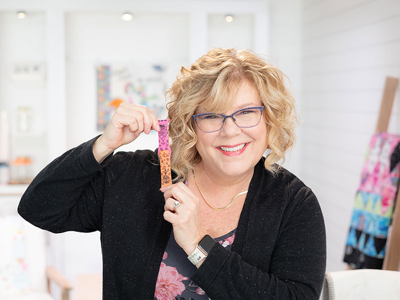 photo of Hope Yoder, smiling and holding a bright orange and pink watch band, in a white room with softly focused embroidered pieces in the distance